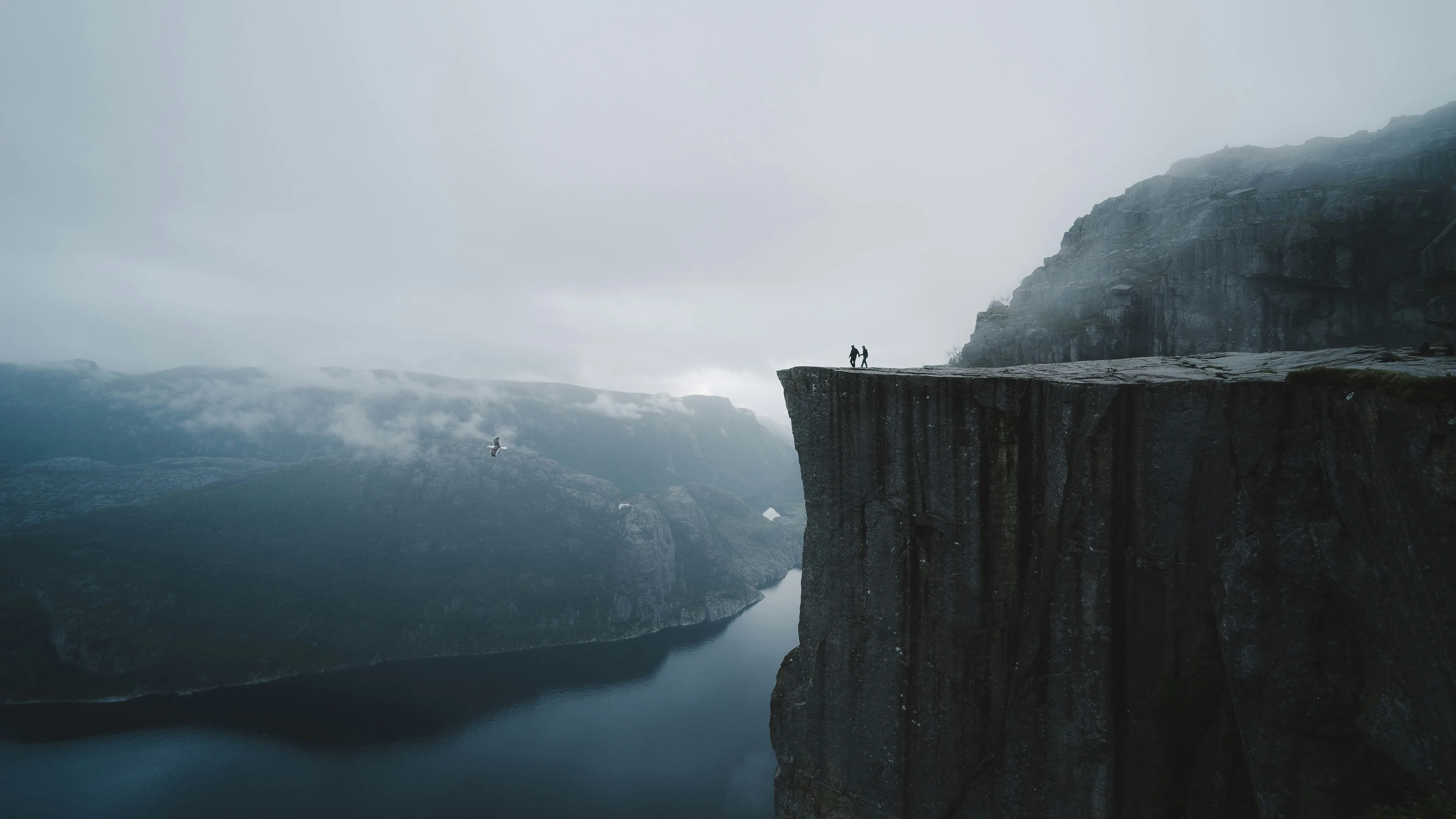 Two people standing on the edge of a dramatic Norwegian cliff overlooking a fjord