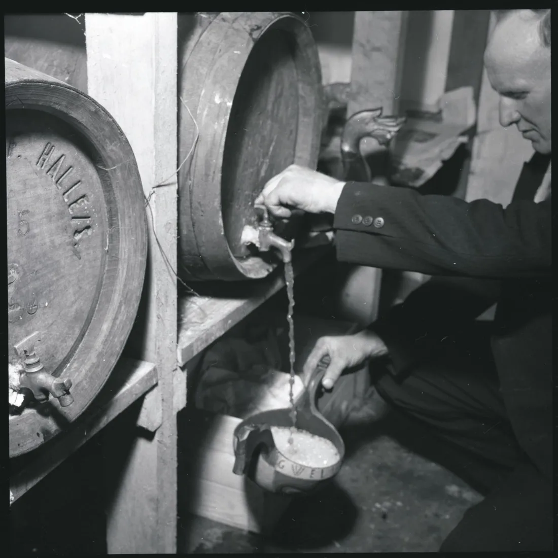 Black and white photograph from 1954 showing a man pouring traditional Norwegian farm beer from a wooden barrel