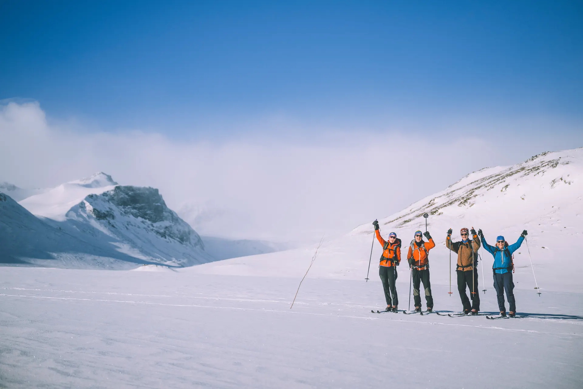 Four skiers celebrating with raised arms in a vast snow-covered mountain landscape under blue skies