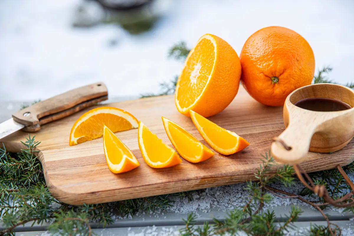 Fresh oranges sliced on a wooden cutting board in the snow, with a traditional wooden cup of coffee and evergreen branches