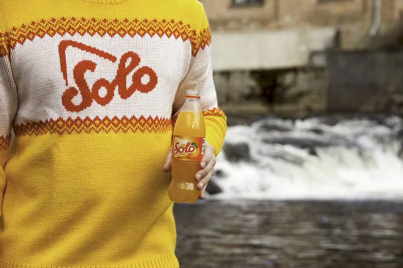 Person wearing a yellow and white knitted sweater with the Solo logo, holding a bottle of Solo orange soda by a river