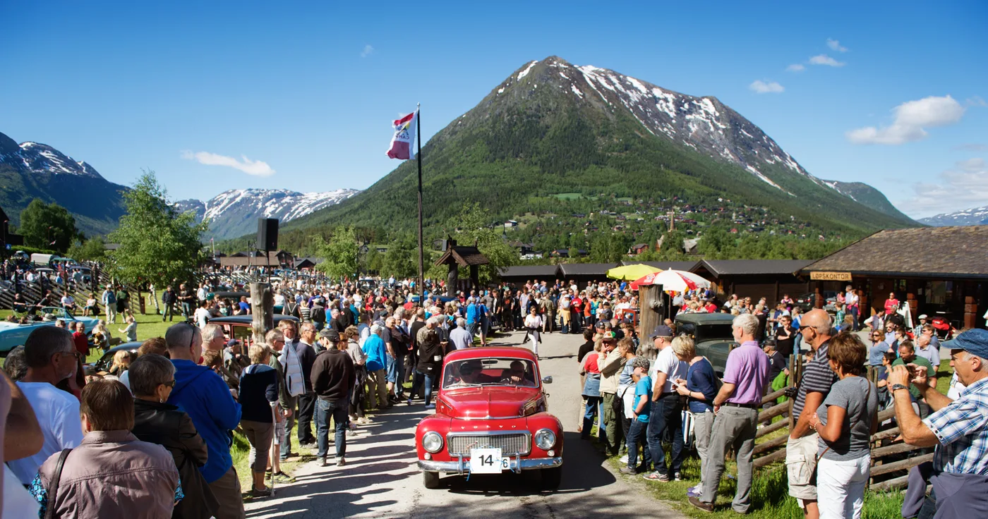 A red vintage Volvo leads a parade through cheering crowds at the Flåklypa Grand Prix in Lom, with dramatic mountain scenery in the background