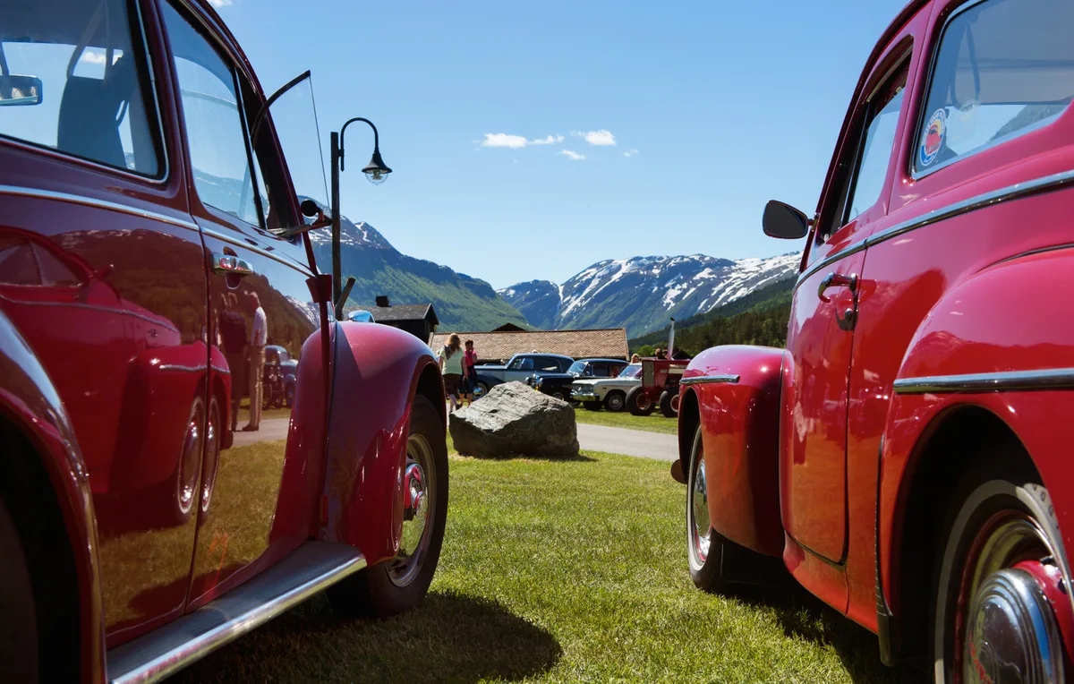 Two red vintage cars frame a scenic view of snow-capped mountains and classic vehicles at the Flåklypa Grand Prix rally in Lom