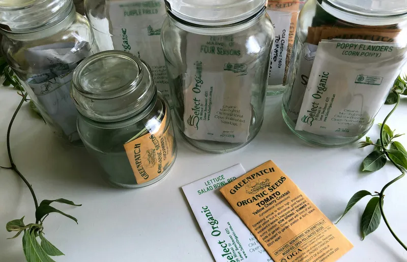 Glass jars and organic seed packets arranged on a table with fresh green cuttings