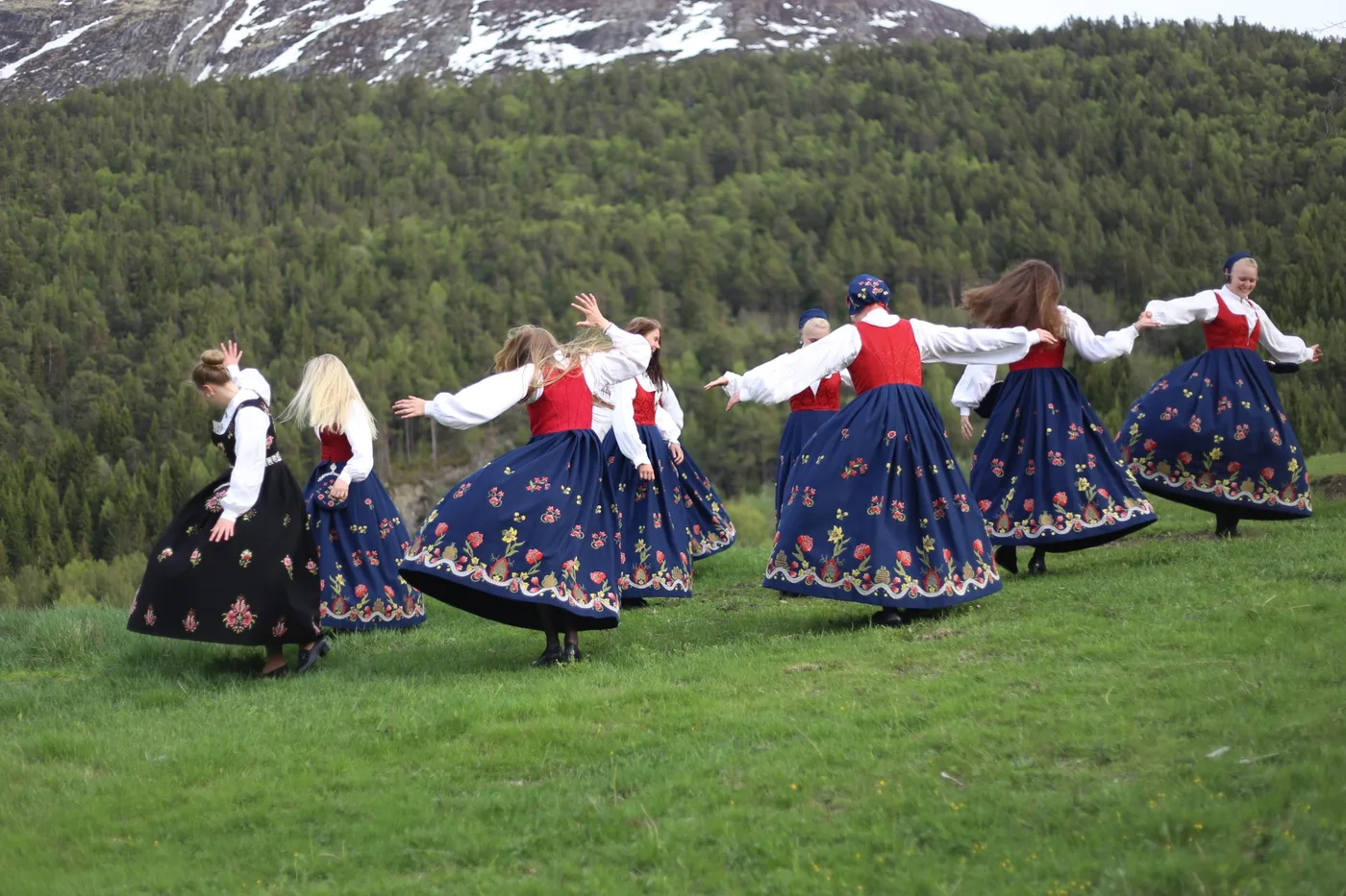 A group of women wearing Graffer bunads hold hands and dance in a circle on a grassy field, with forested mountains in the background