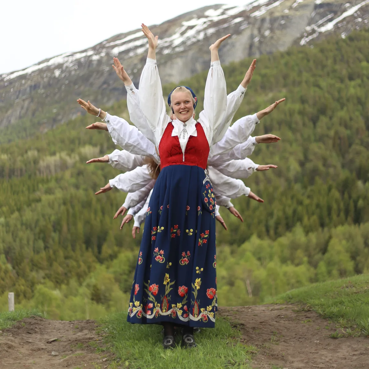 A woman in a Graffer bunad stands smiling outdoors with multiple extended arms radiating behind her in a performance art composition, with snow-capped mountains in the background