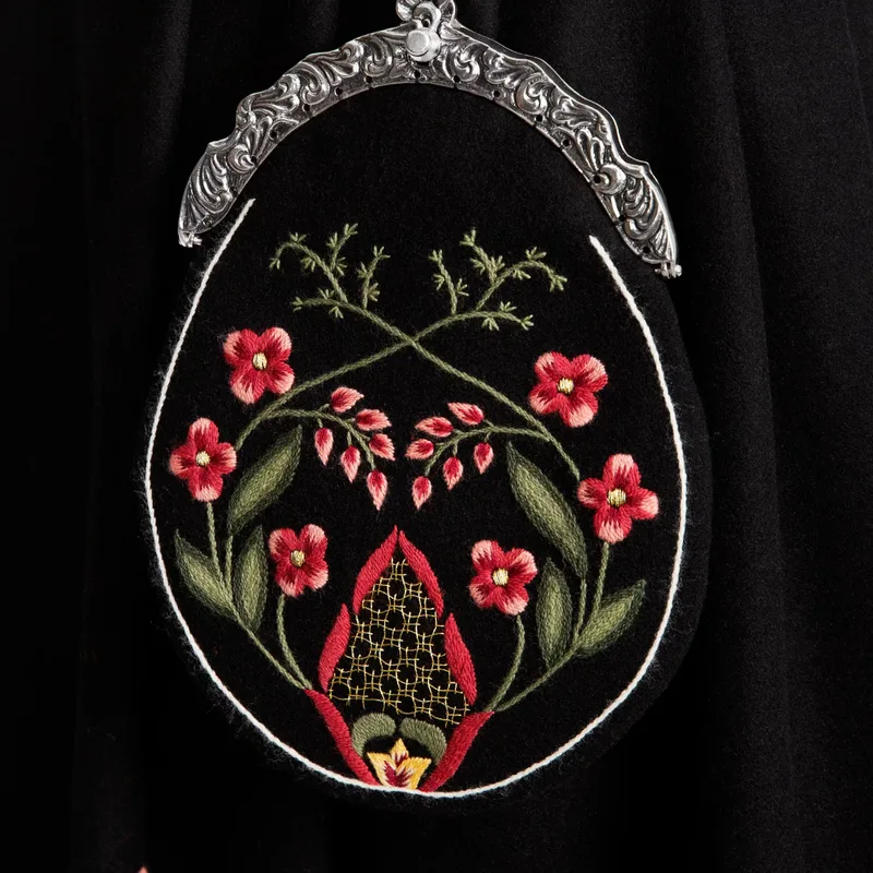 Close-up of a detachable bunad pocket in black wool, richly embroidered with pink and red floral motifs, green leaves, and a silver clasp frame