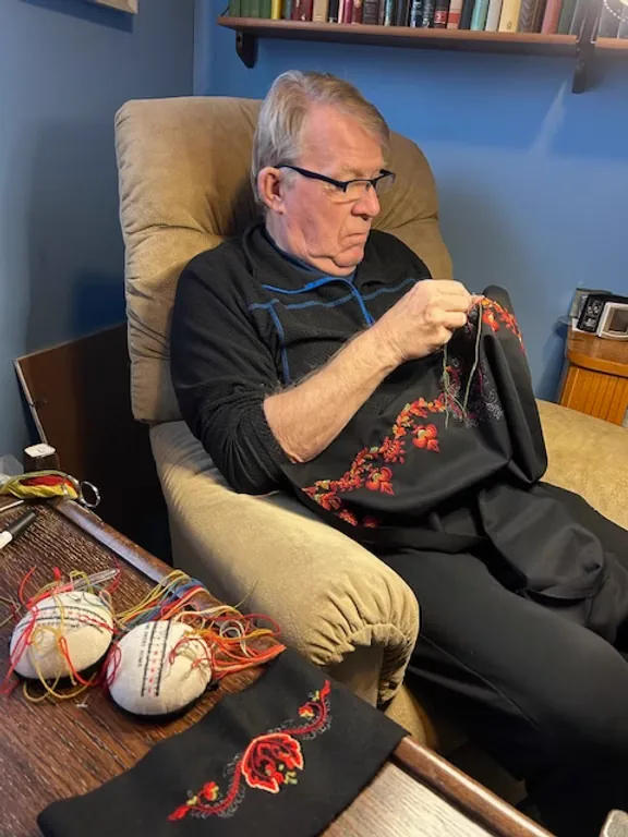 Eighty-year-old Alf 'Beppe' embroidering a black bunad bodice with red wool embroidery in his armchair, surrounded by colourful threads