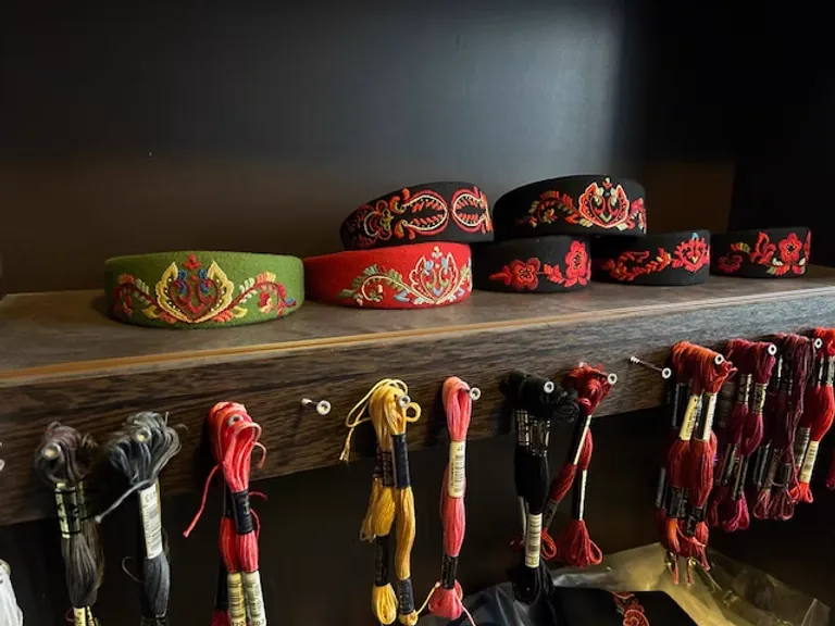 Display of raudsaum-embroidered bunad headbands in green, red, and black on a wooden shelf, with colourful embroidery threads hanging beneath