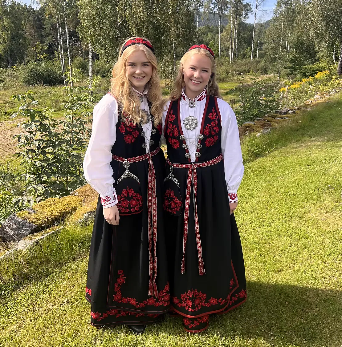 Sisters Leah and Tine Sofie Thorstensen wearing matching Vest-Telemark Raudsaum bunads in a Telemark garden with birch trees