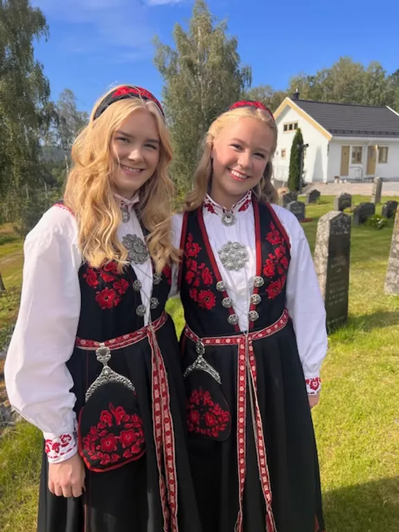 Sisters Leah and Tine Sofie Thorstensen in Vest-Telemark Raudsaum bunads in front of a white wooden church in Telemark