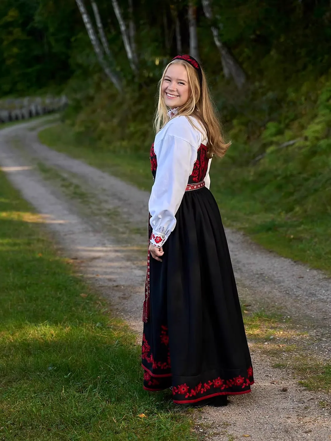 A young woman in a Vest-Telemark Raudsaum bunad walking on a forest path in evening light, looking back over her shoulder