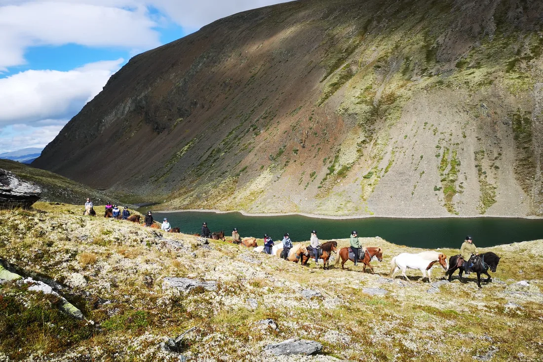 A line of riders on Icelandic horses trekking along a mountain lake with steep cliffs rising behind them