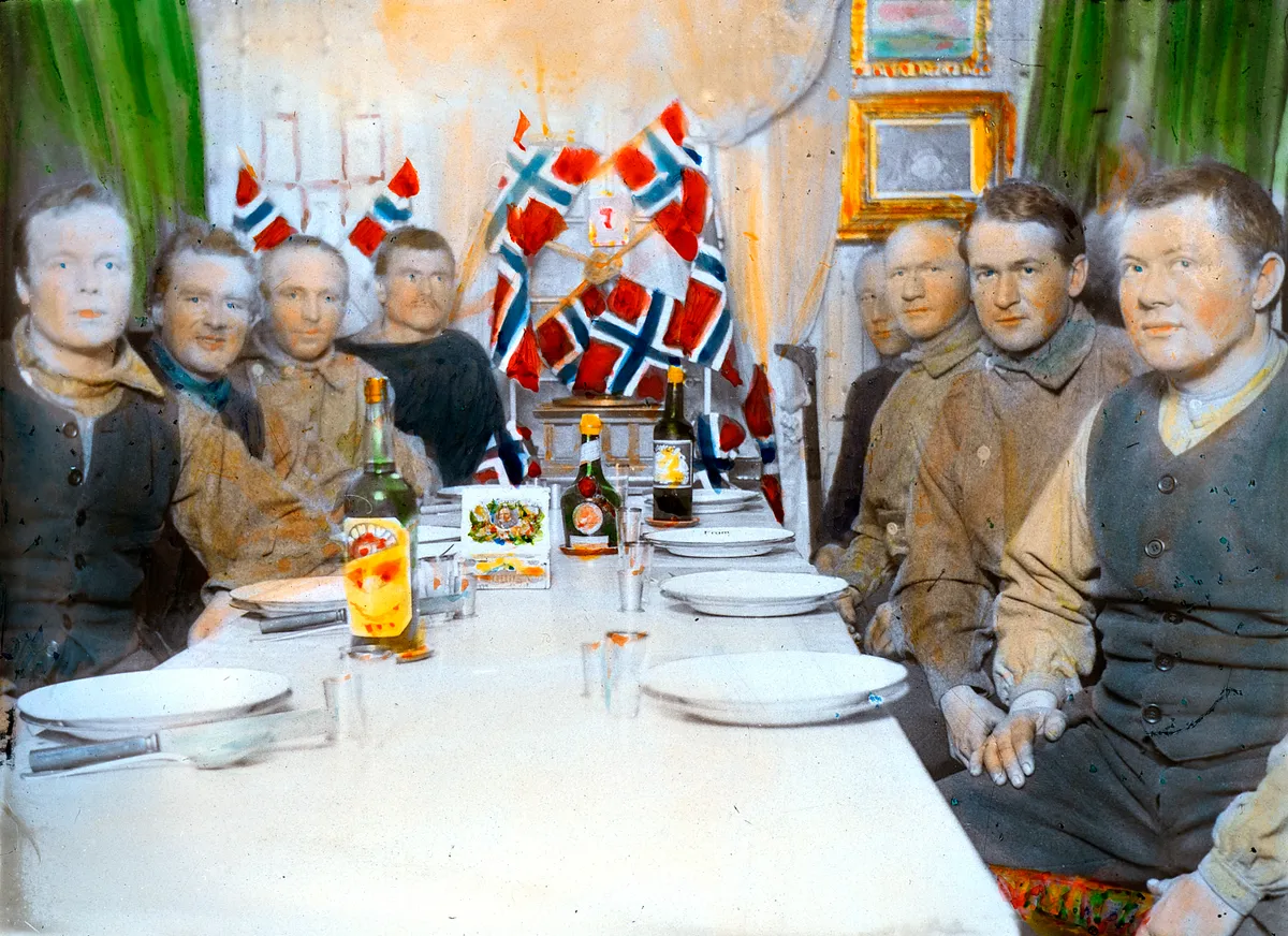 Historic photograph from 1912 showing a formal dinner in Whale Bay, Antarctica, with aquavit on the table – Roald Amundsen is among the guests