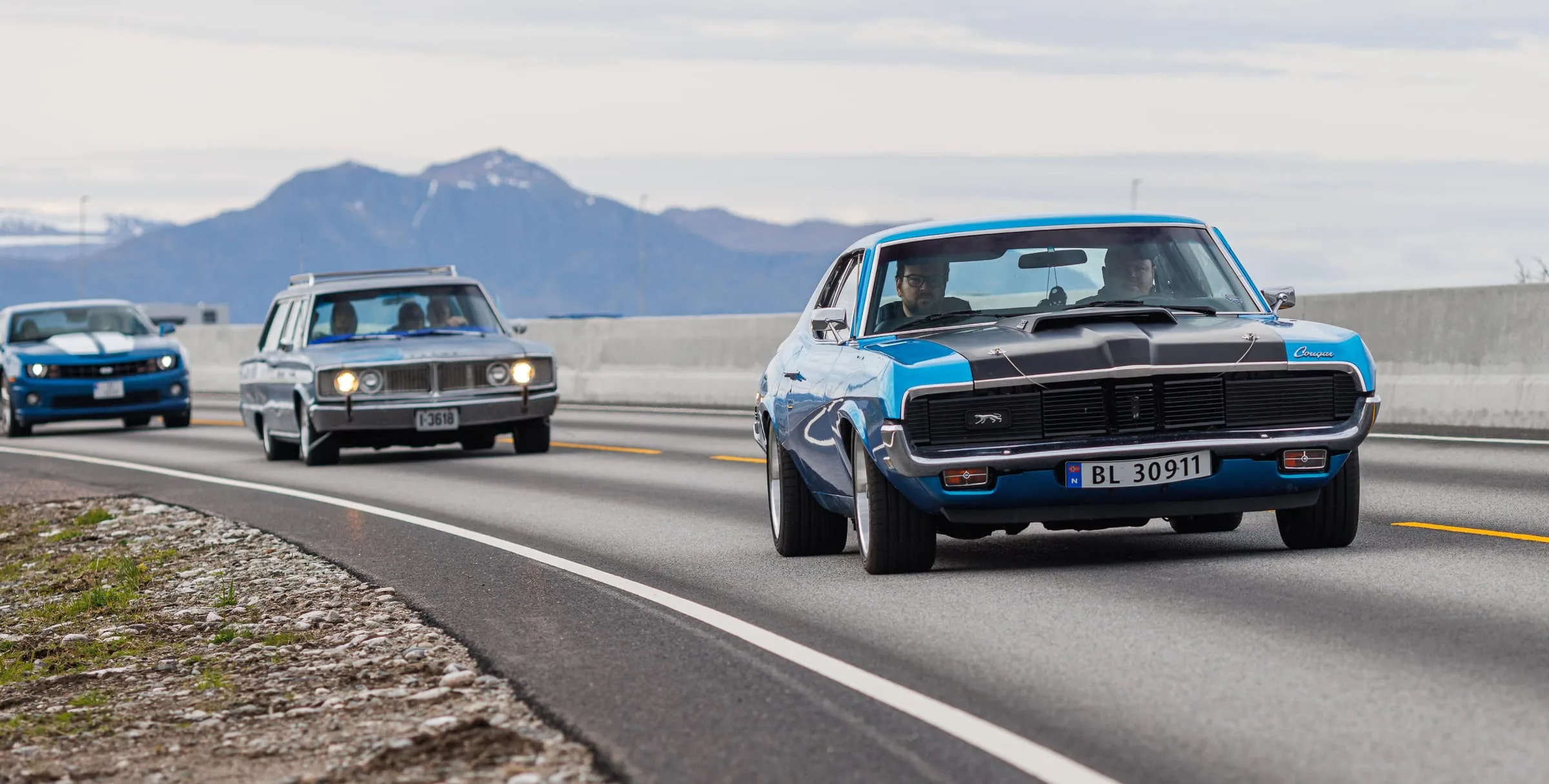Classic American muscle cars from Seaside Custom driving on Nordøyvegen with mountains in the background
