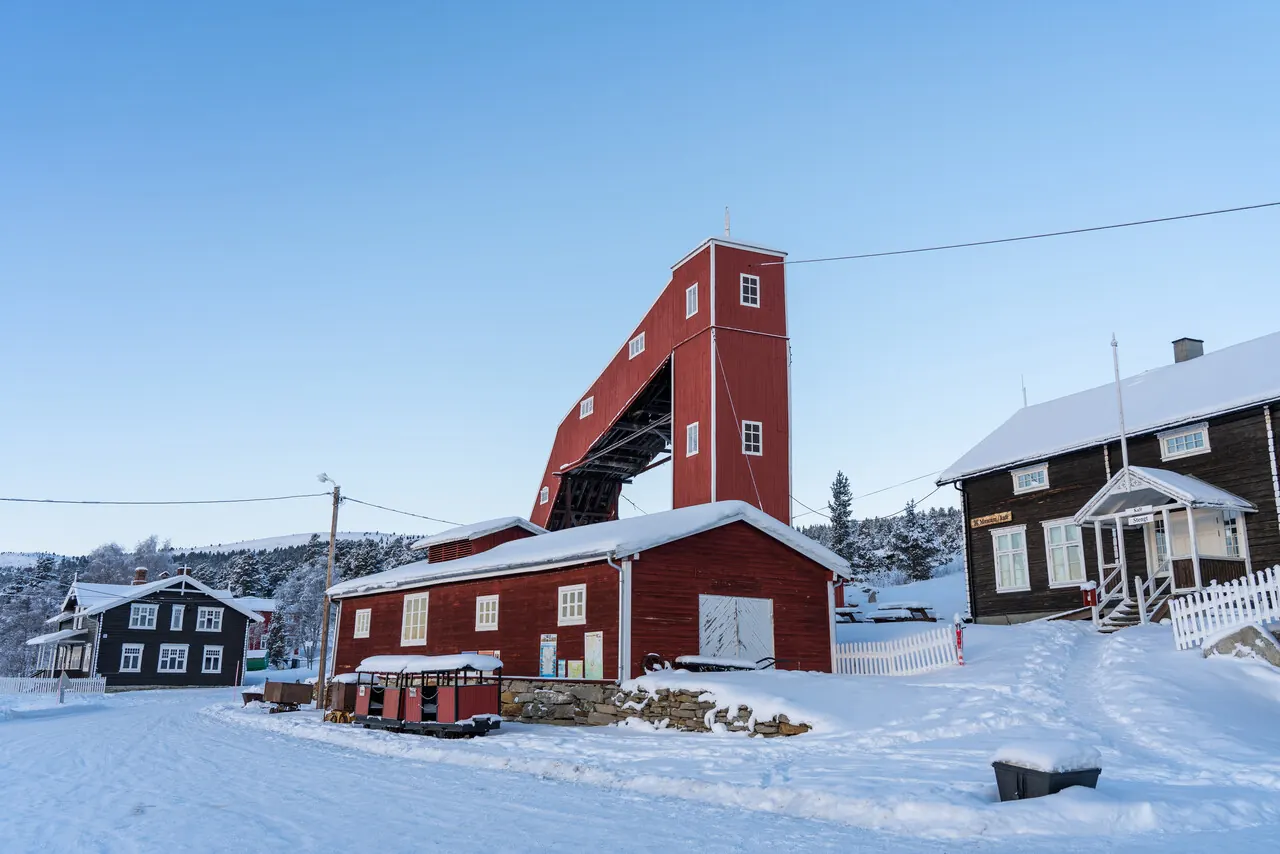 Red timber mine headframe and buildings at Folldal Verk in winter, with snow-covered ground under a clear sky