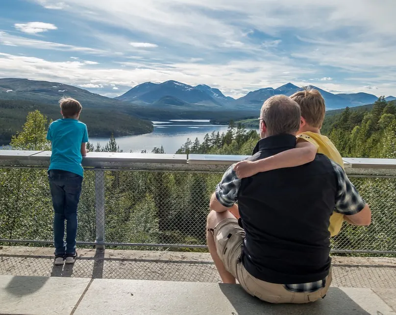 An adult and two children standing at the Sohlbergplassen viewpoint, gazing over a forested valley, lake, and the Rondane mountain range