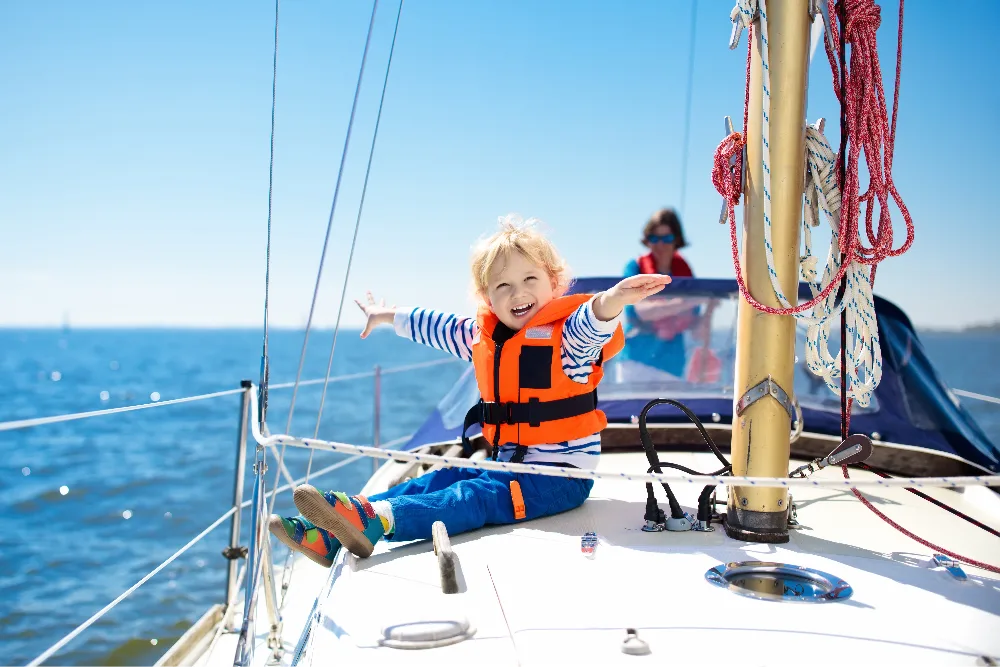 A child wearing an orange life jacket on a sailboat, enjoying a sunny day at sea
