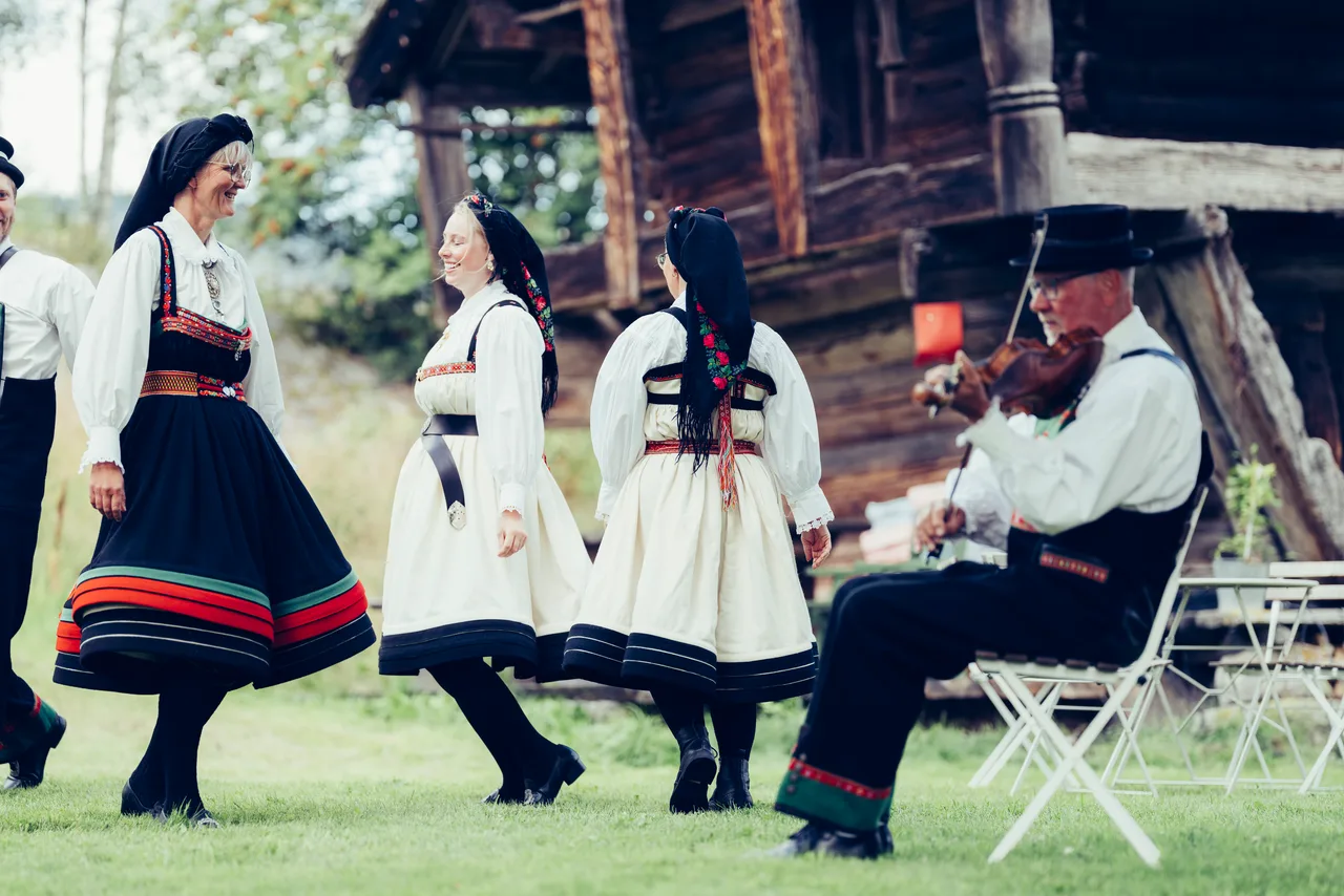 Two couples in Setesdal bunad dancing on green grass while a fiddler accompanies them beside a wooden storehouse