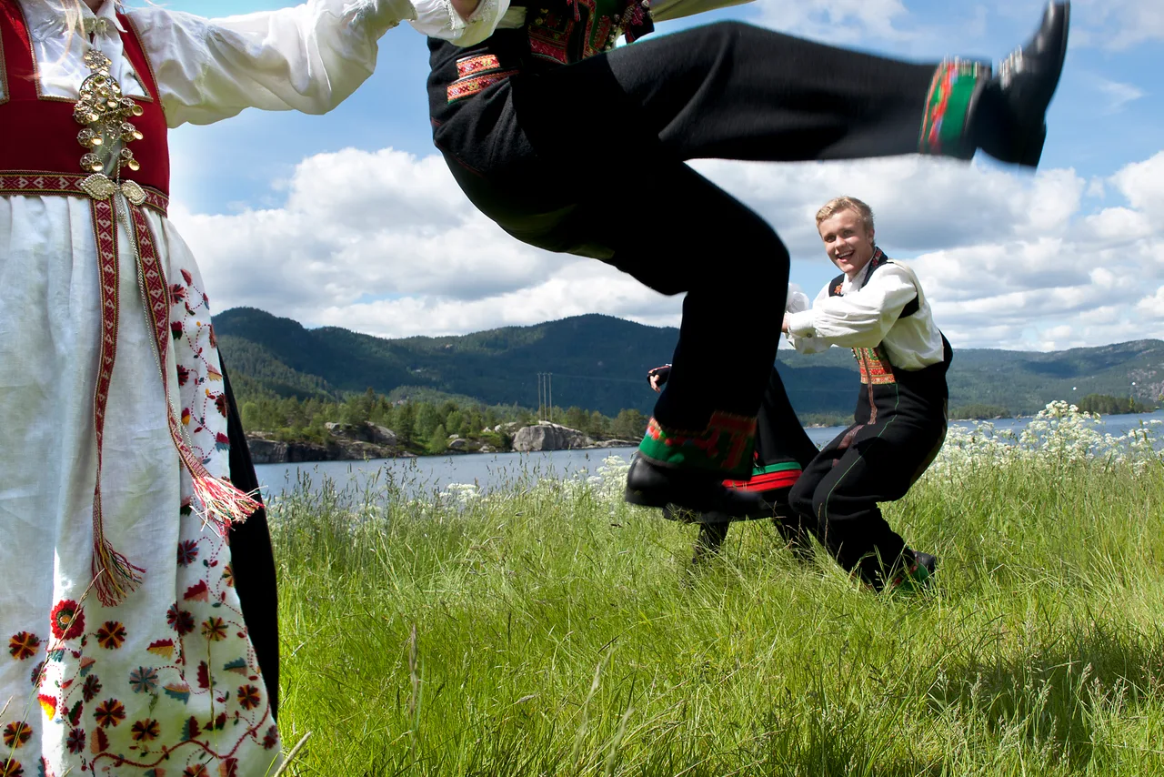 A dancer in bunad leaping high into the air during an athletic sprekleik dance, with mountains and a lake in the background