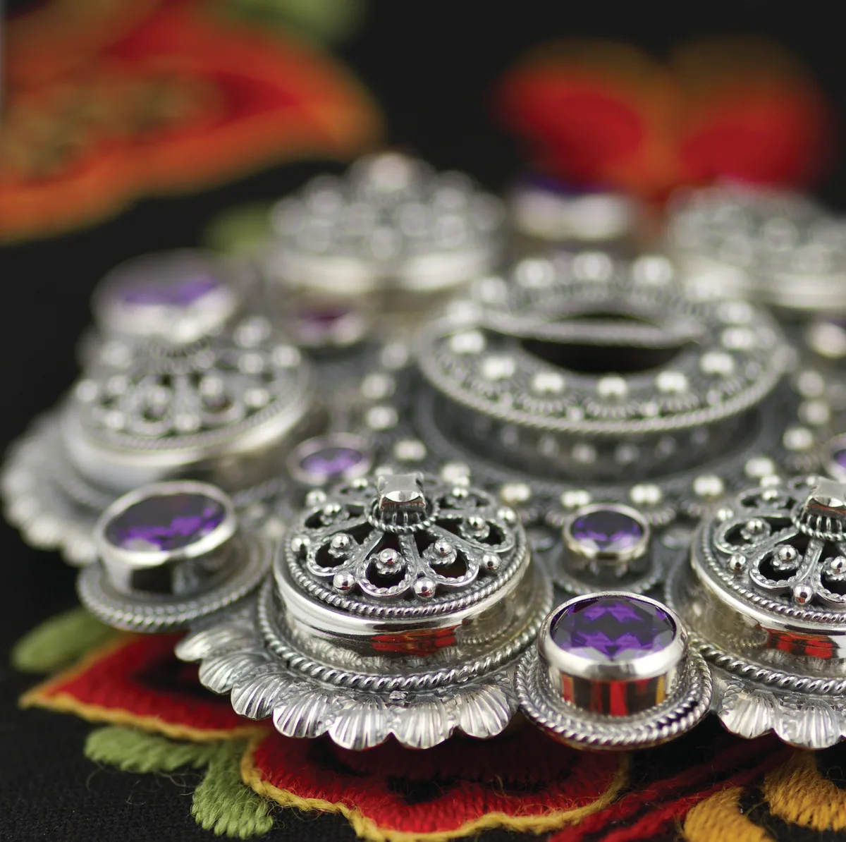 A stunning bolesølje brooch in polished silver with deep purple gemstones, resting on colourful embroidered bunad fabric