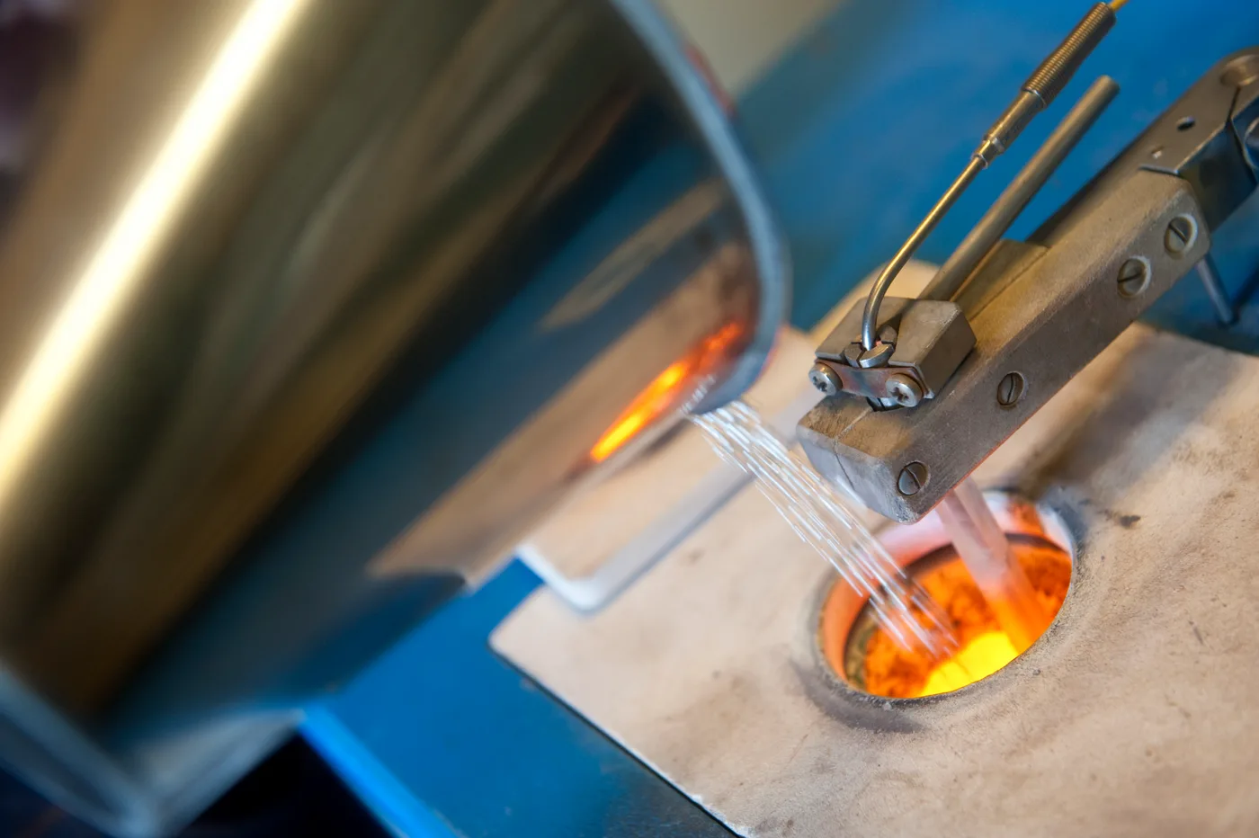 Close-up of a silversmith’s workshop showing silver wire being drawn through a die, with the glow of a furnace in the background