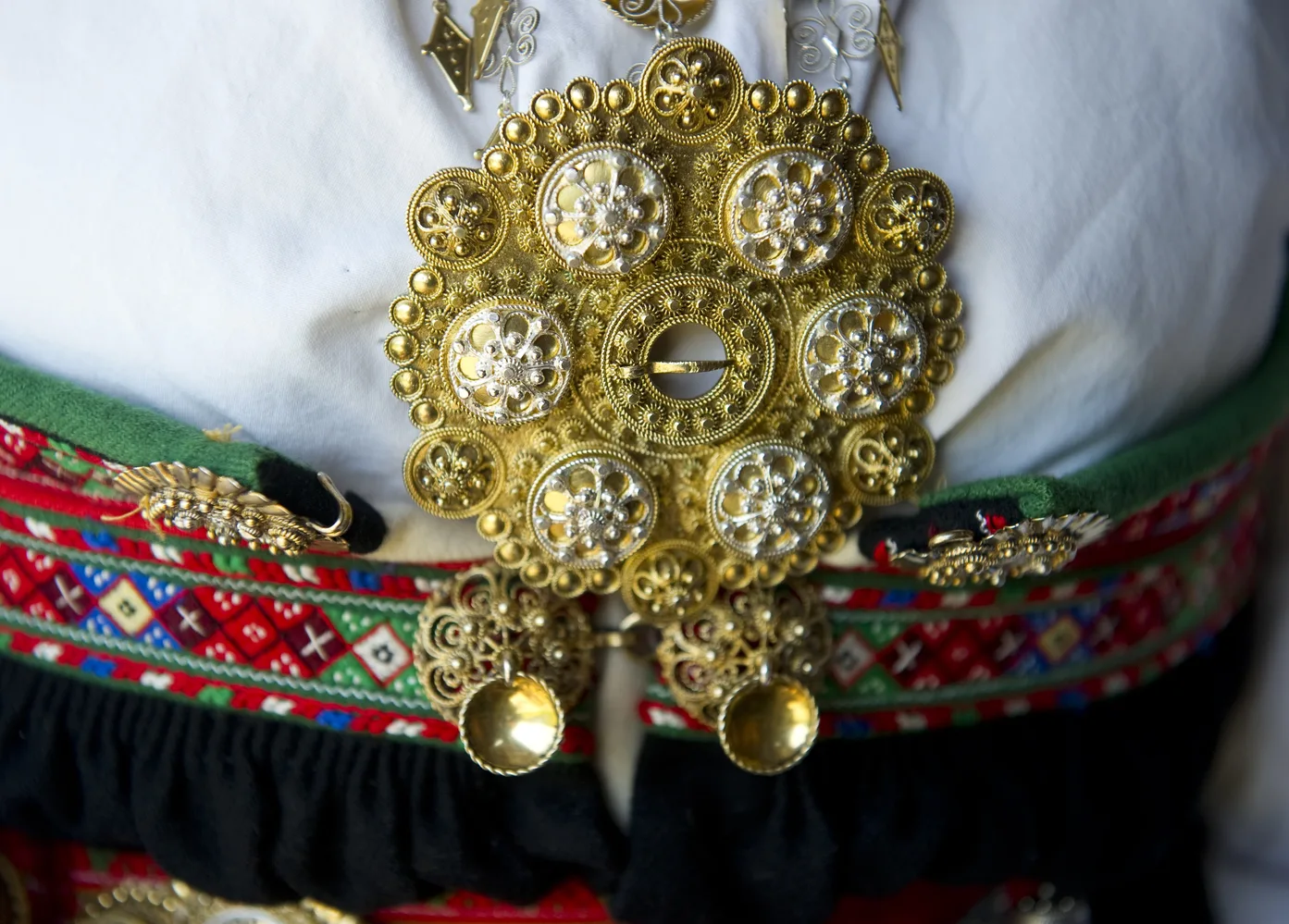 A gilded sølje brooch with filigree flower patterns worn on a Setesdal bunad, with colourful woven belt visible beneath