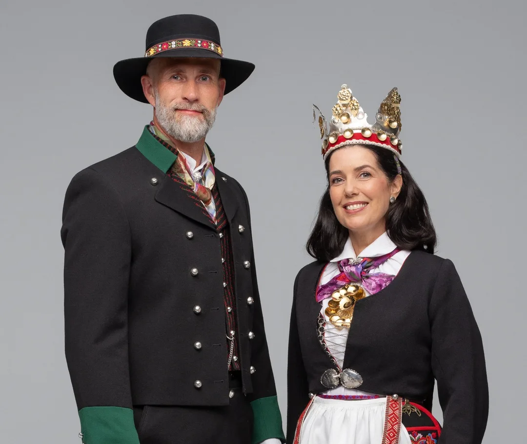 A couple in Vest-Agder bridal attire – the bride wears a bunad with a silver crown and white apron, the groom in a black jacket with silver buttons and traditional hat