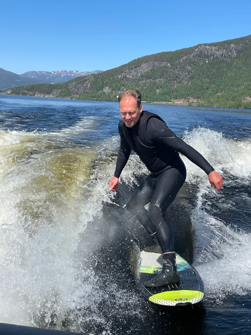 A wakesurfer riding freely behind a boat on lake Krøderen, surrounded by green hillsides