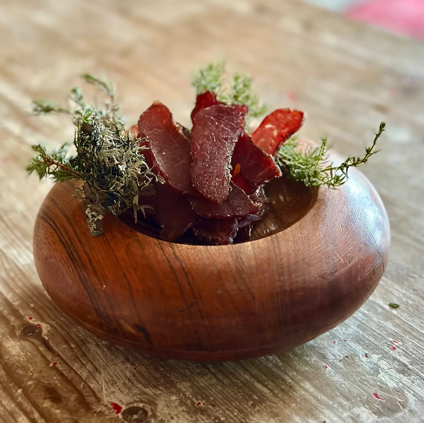 Thin slices of cured wool pig meat served in a handcrafted wooden bowl, garnished with sprigs of juniper and moss