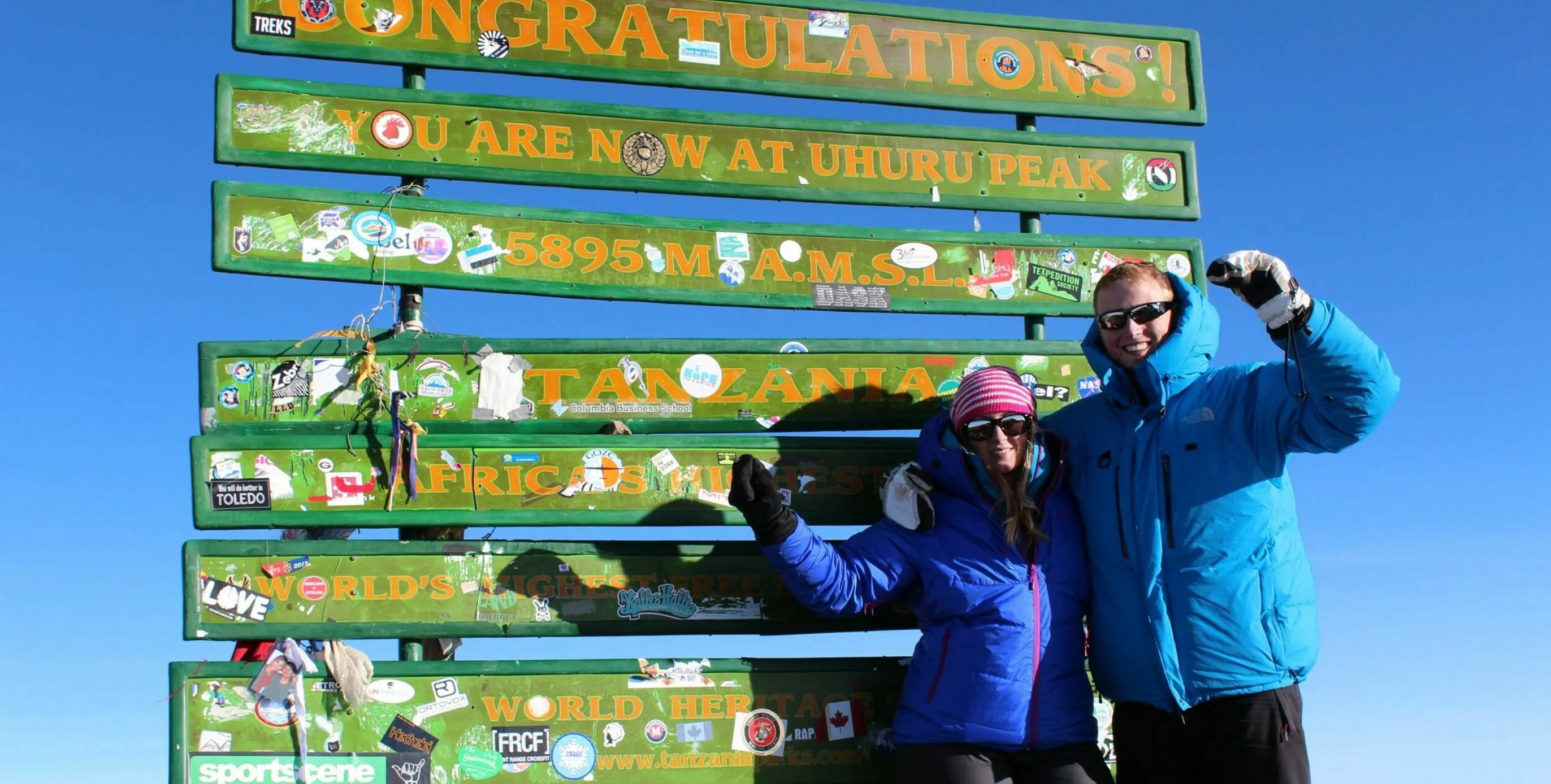 Two people in blue jackets celebrating at the Uhuru Peak summit sign on Mount Kilimanjaro, 5895 metres above sea level.
