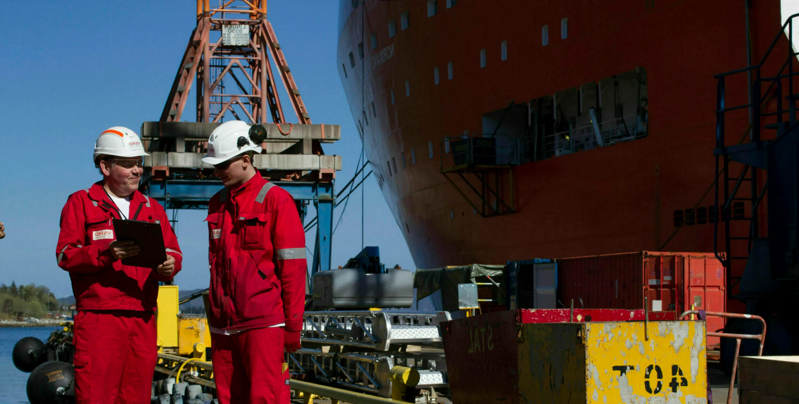 Two VARD employees in red coveralls and safety helmets go through a checklist on the quay next to a ship hull under construction.