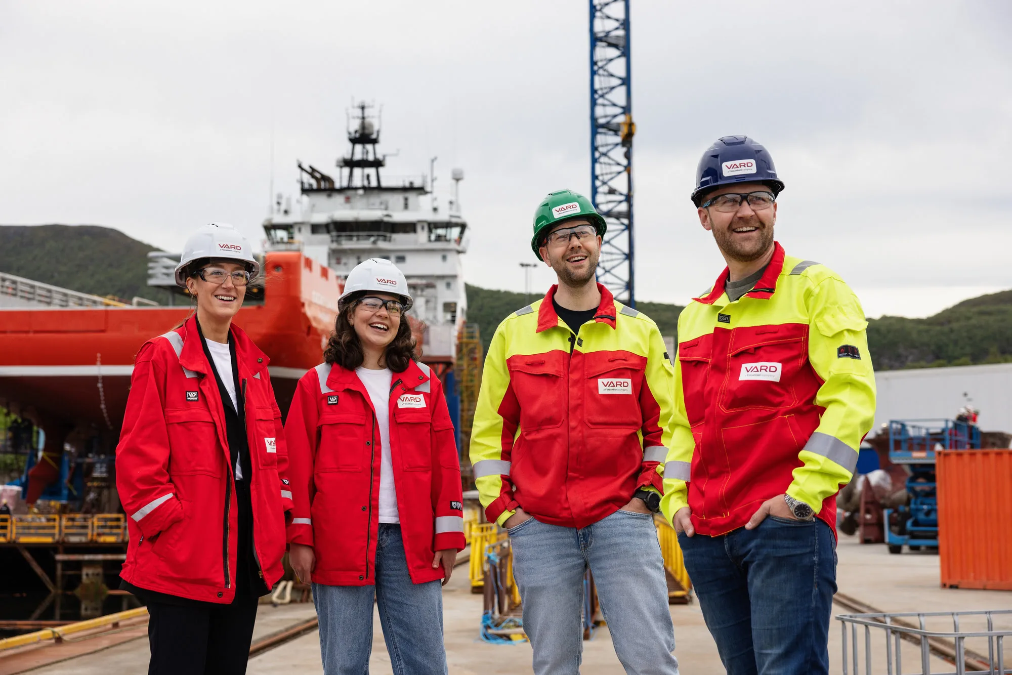 Four smiling VARD employees in red coveralls and hard hats standing at the shipyard with a vessel under construction behind them.