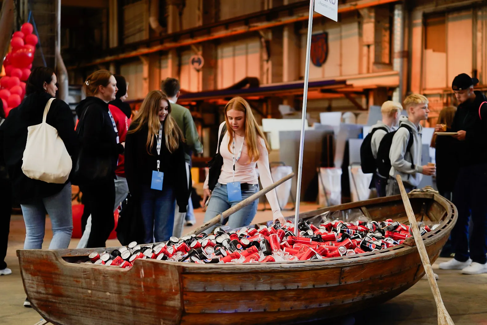 Teenagers at an indoor innovation event gather around a wooden rowing boat filled with red and white drink cans at Vard Langsten.