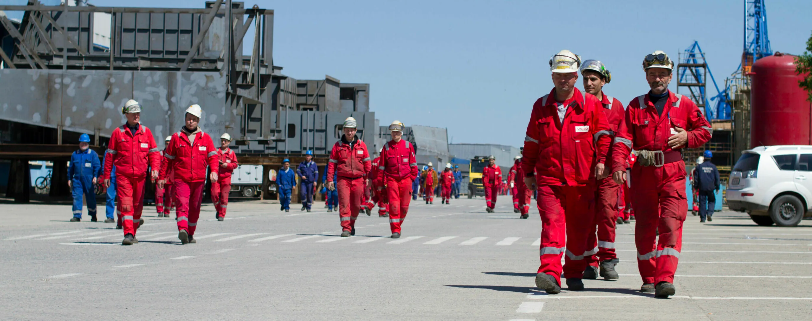 A group of VARD shipyard workers in red coveralls and hard hats walking across the yard, highlighting teamwork and safety.