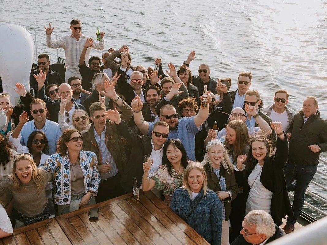 A large group of smiling VARD employees on the deck of a boat at sea, raising their hands and enjoying a social event together.