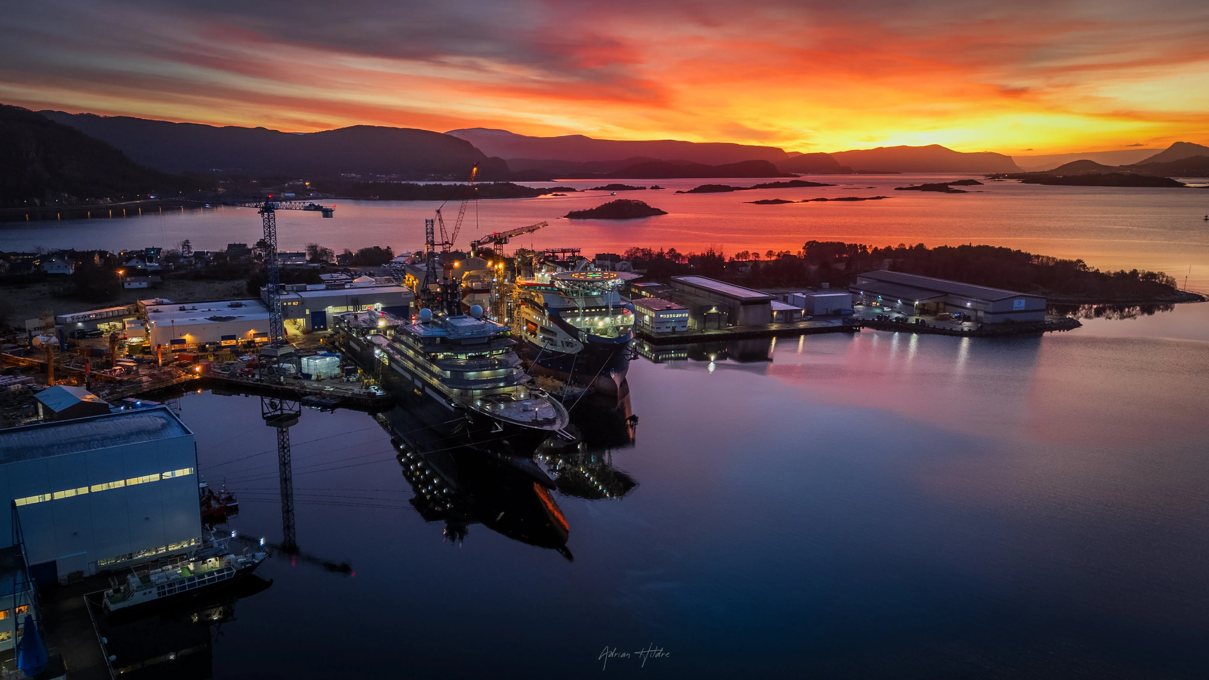 Aerial view of the VARD shipyard on Sunnmøre at sunset, with vessels docked and mountains silhouetted against an orange sky reflected in the fjord.