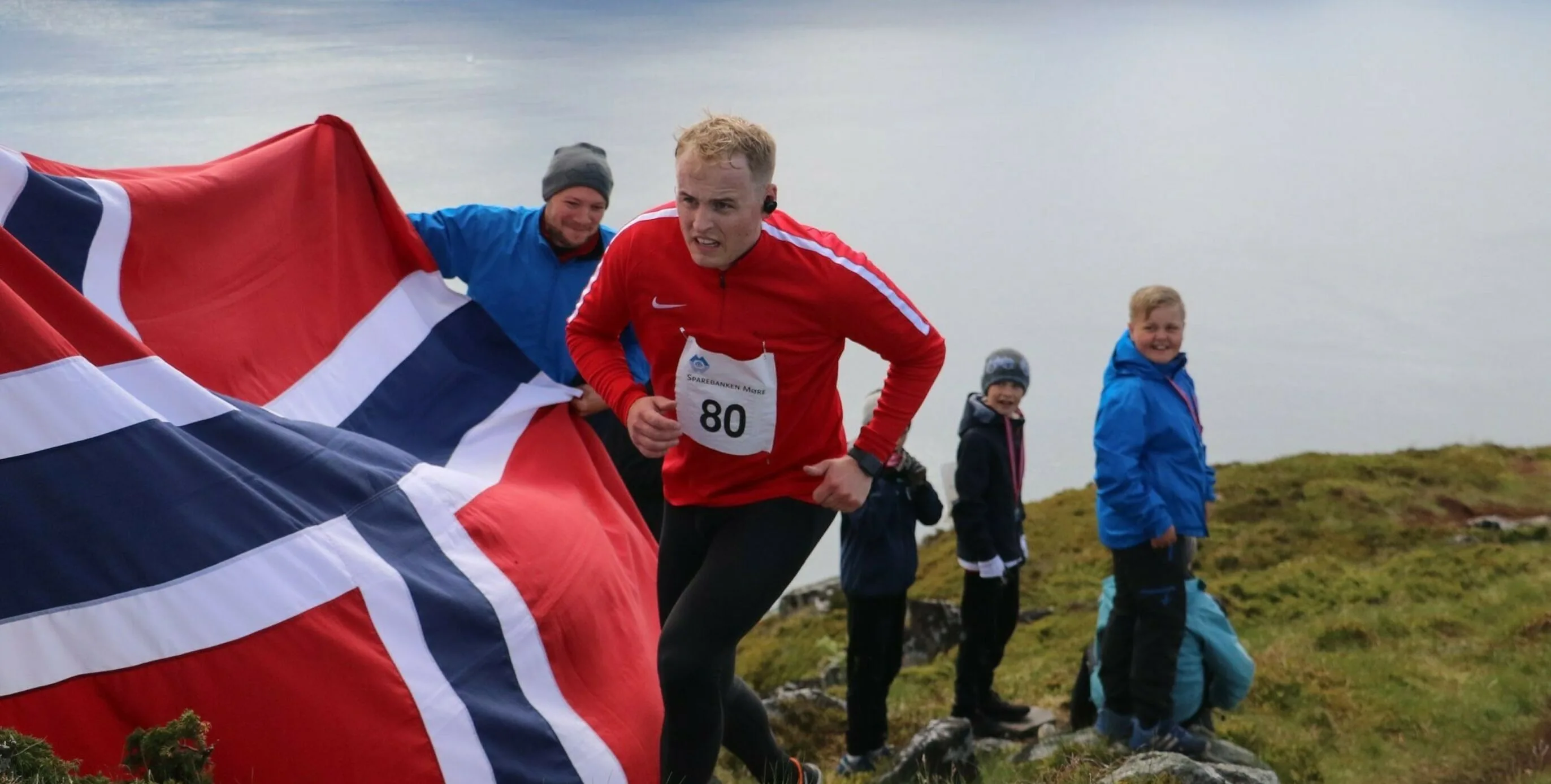 A runner wearing a red jacket and race bib number 80 runs along a windswept coastal hill while another person holds a large Norwegian flag behind him.