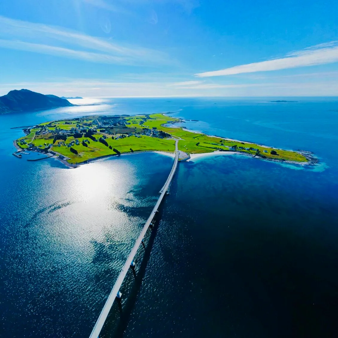 Aerial view of a green coastal island connected by a long bridge, surrounded by deep blue sea under a bright sky.