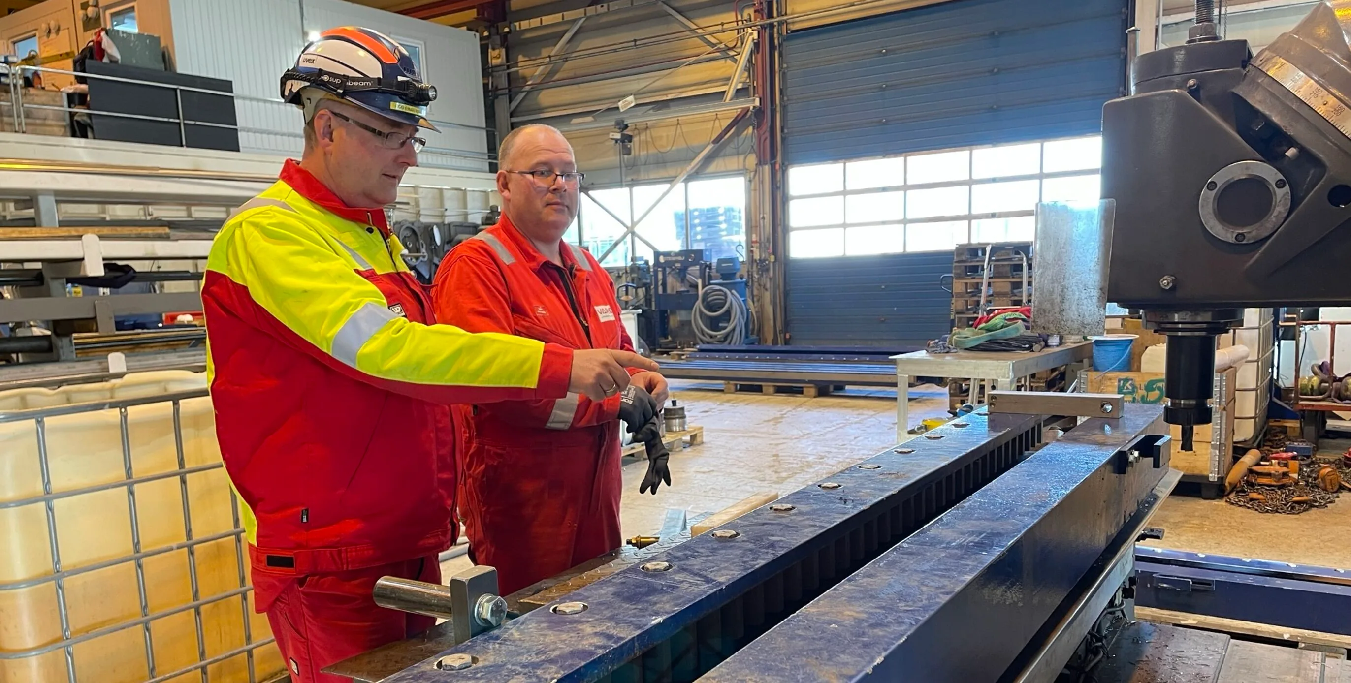 Odd-Einar and a colleague working together on heavy industrial machinery in the VARD workshop hall, with bright daylight from the large doors.
