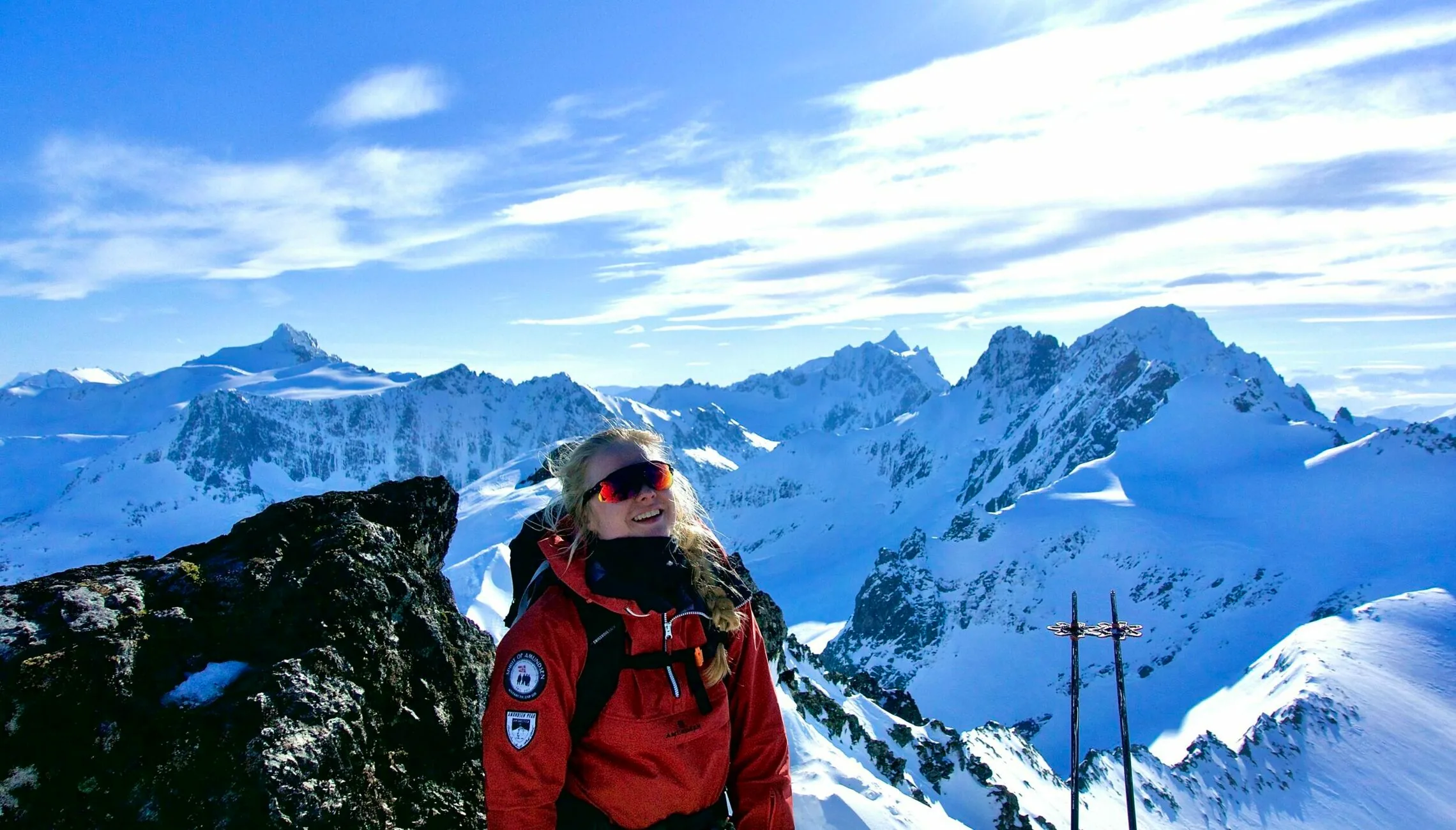 Person in a red jacket standing on a snowy mountain viewpoint with dramatic alpine peaks and a blue sky in the background.
