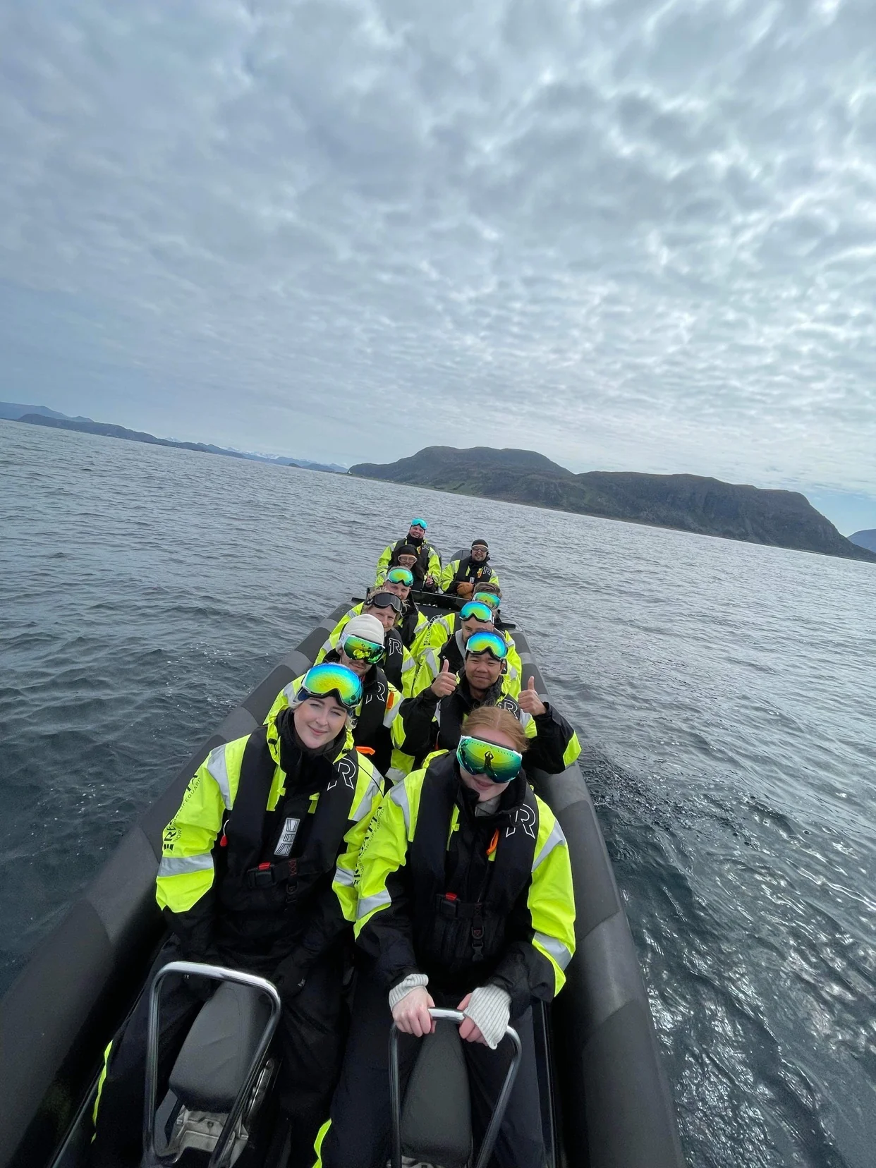 A group of VARD employees in bright survival suits sitting close together in a RIB boat on the fjord, with mountains in the background.