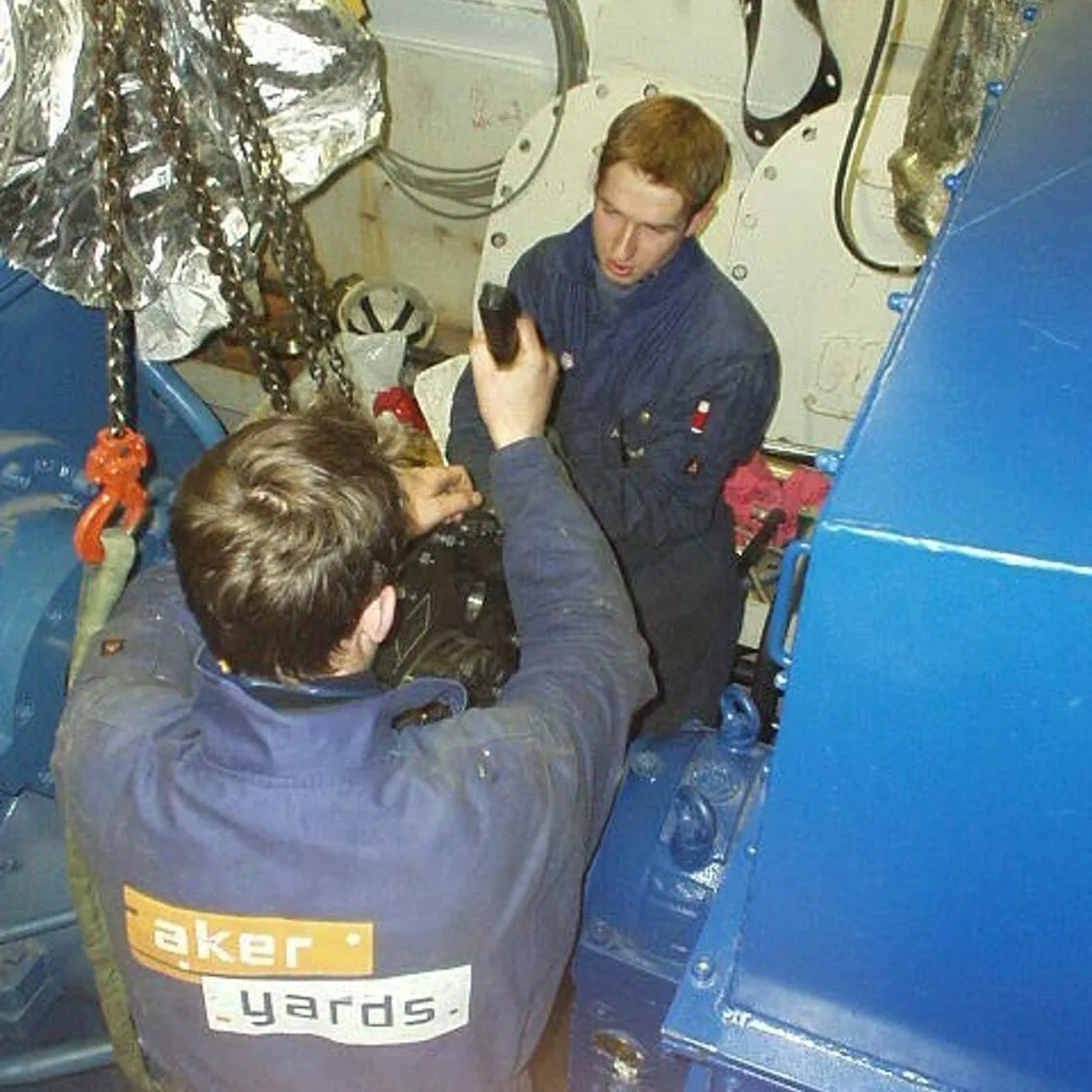 Two young mechanics in Aker Yards overalls working on heavy machinery in a ship’s engine room, circa 2000.