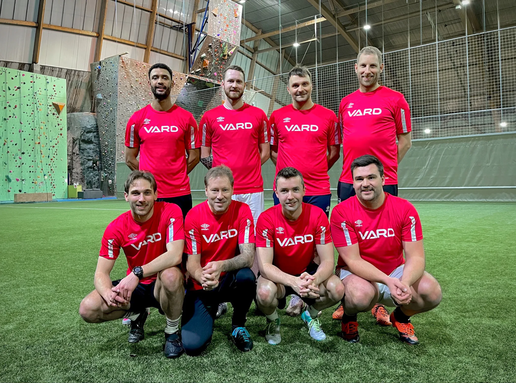 Eight VARD employees in red VARD football shirts posing for a team photo on an indoor football pitch.