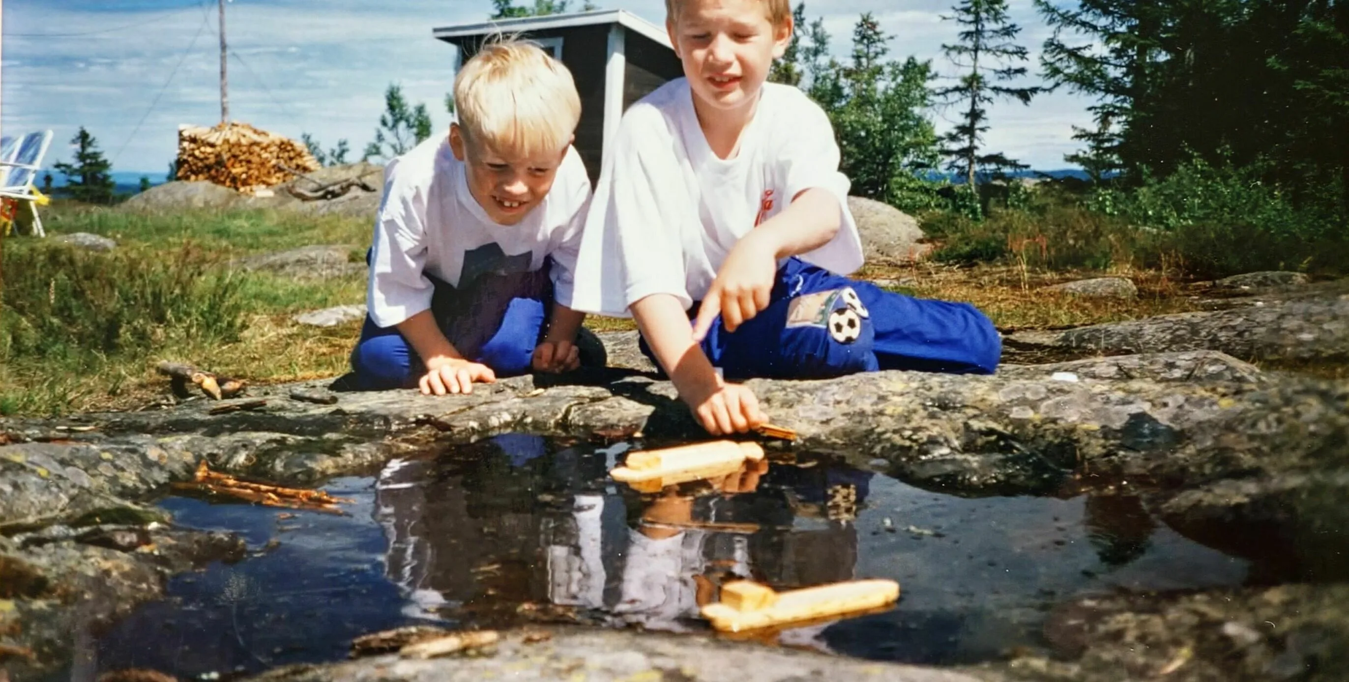 Two young boys kneel on sunlit rocks outdoors, watching small wooden boats float in a shallow puddle, with trees and a cabin in the background.