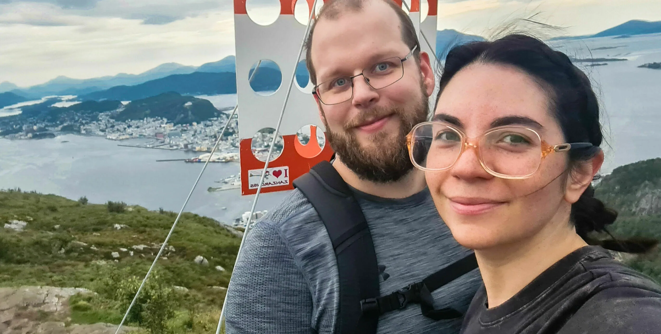 Gema with a companion taking a selfie on a mountaintop above Ålesund, with fjords and islands in the background.