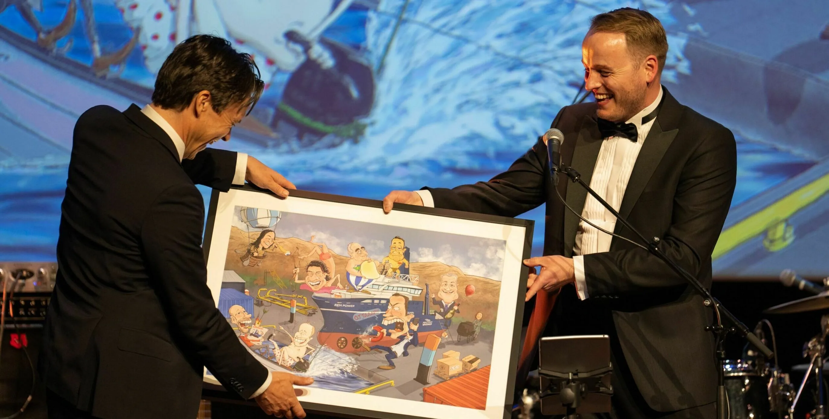 Two men in tuxedos smile on a stage during an event while presenting a large framed illustration; a microphone stand and a projected background image are visible behind them.