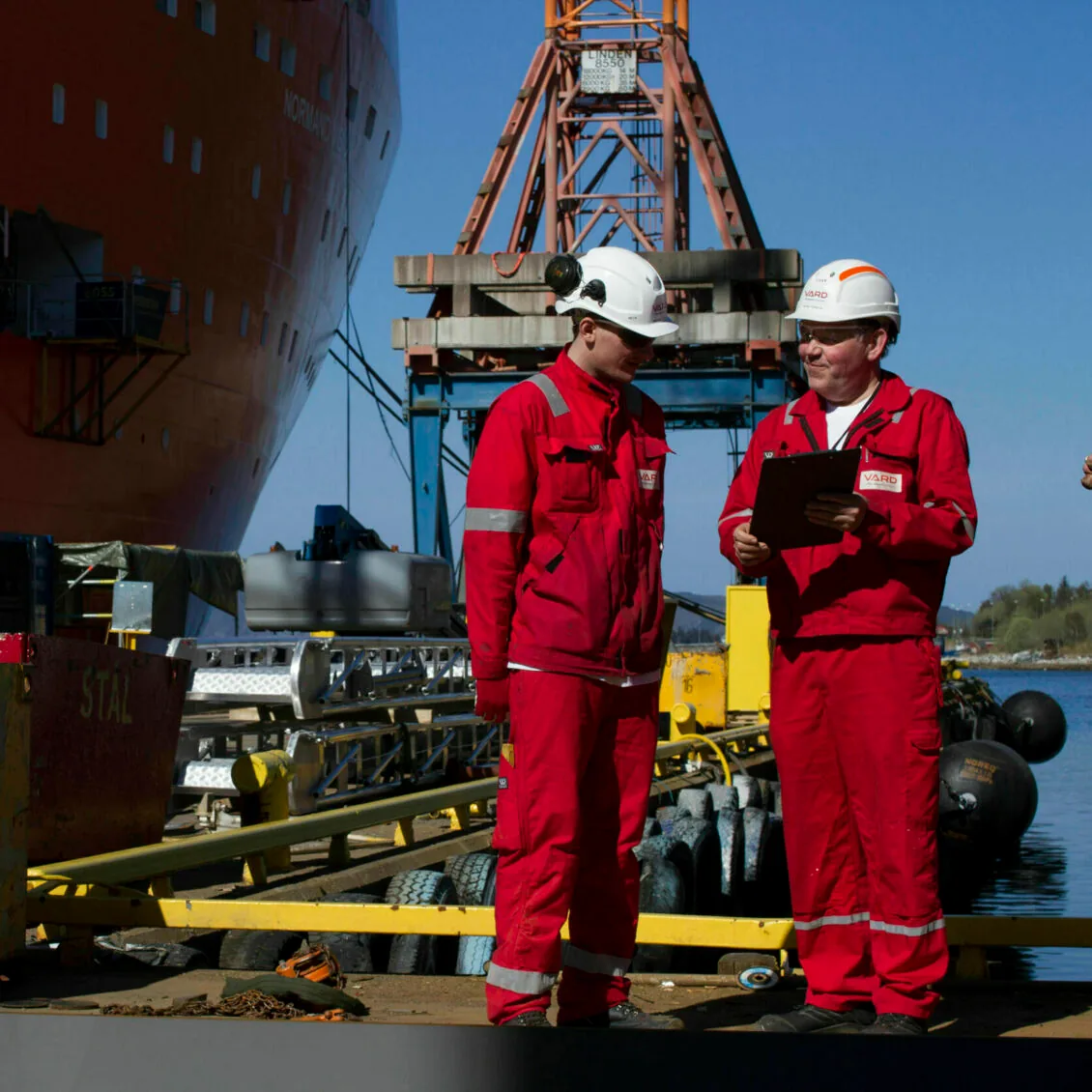 Two VARD employees in red coveralls and safety helmets stand on the quay with a ship and crane behind them, reviewing a checklist.