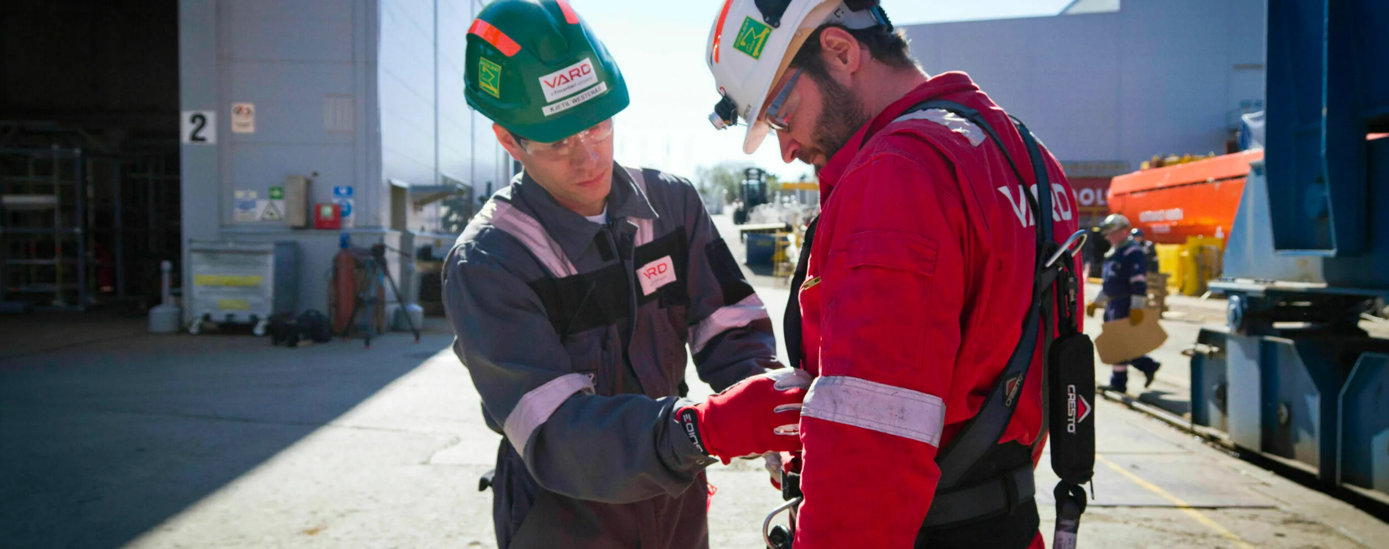 A VARD colleague helps a co-worker fasten a safety harness on the shipyard, sunlight catching their helmets and high-visibility gear.