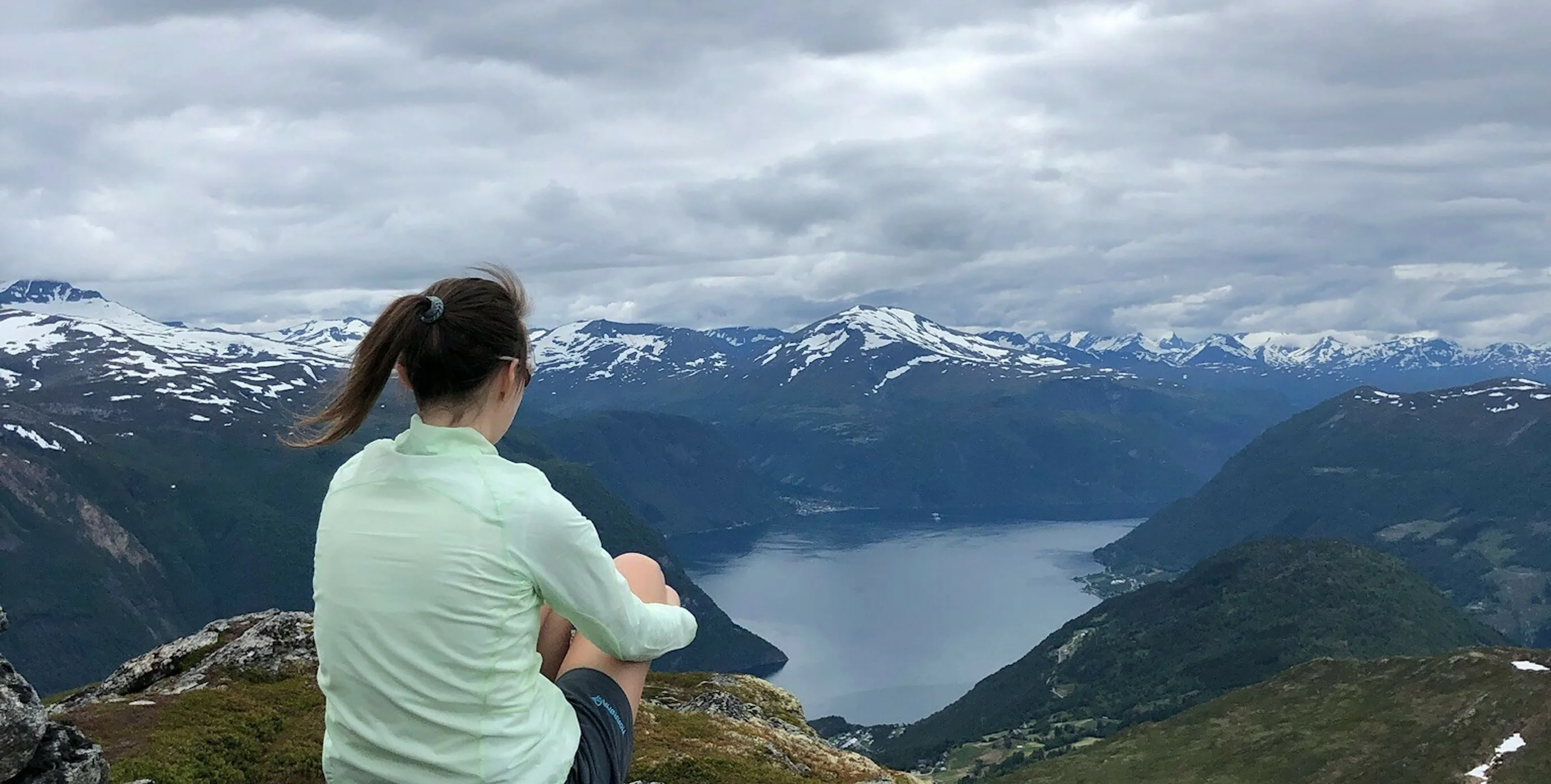 A woman sits on a mountain ridge overlooking a fjord and snow-capped peaks in Sunnmøre.
